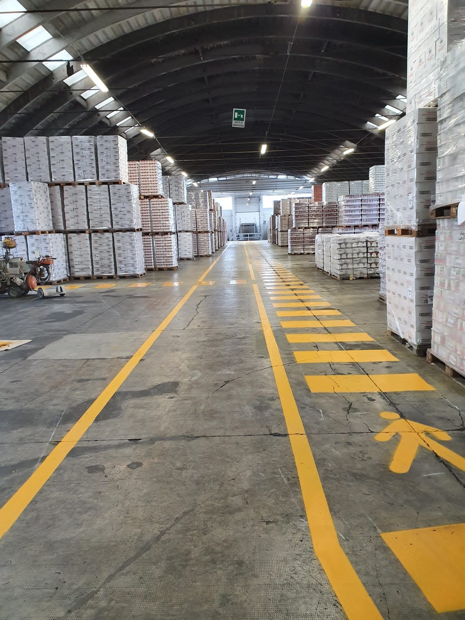 Un ampio corridoio di magazzino scarsamente illuminato, con alte pile di scatole bianche pallettizzate su entrambi i lati e un percorso pedonale dipinto.