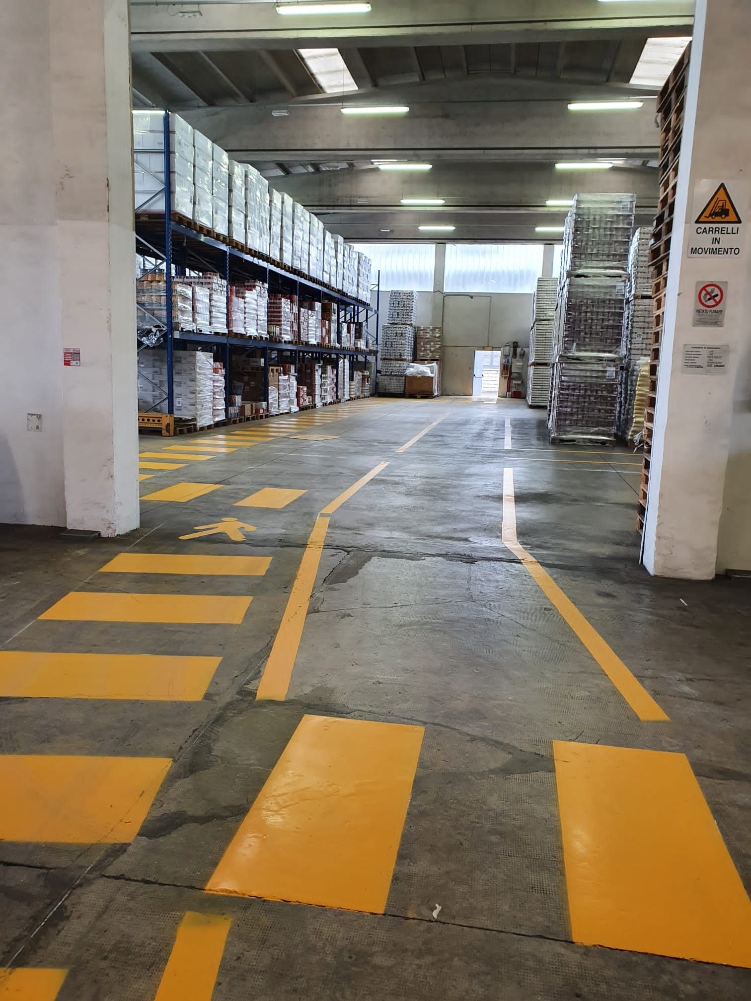 L'interno di un magazzino presenta alte scaffalature metalliche sulla sinistra e attraversamenti pedonali dipinti di giallo sul pavimento.