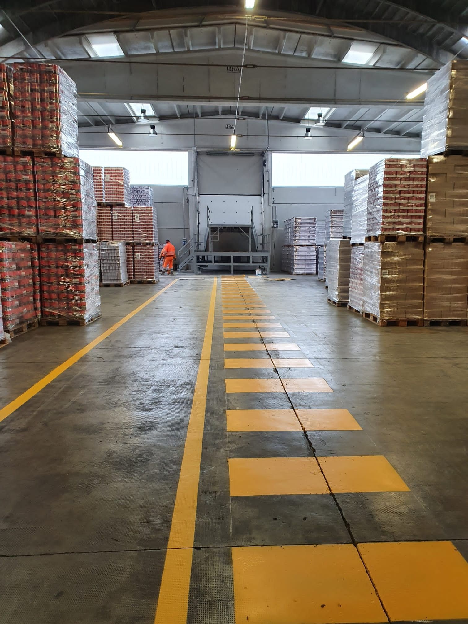 Un corridoio di magazzino con pile di merci pallettizzate su entrambi i lati, delimitato da linee gialle sul pavimento che conducono a una baia di carico.