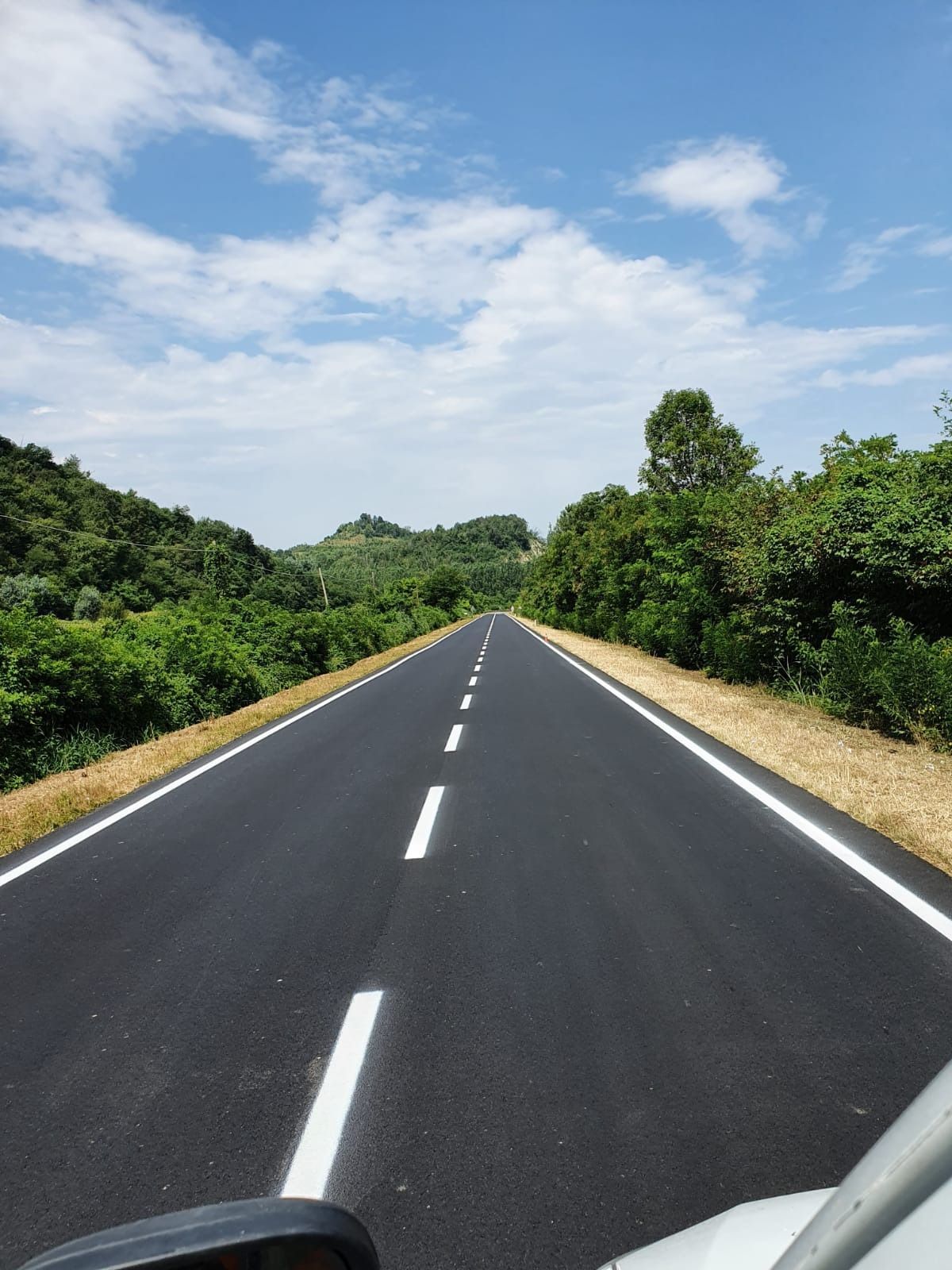 Una strada appena asfaltata si estende a perdita d'occhio, fiancheggiata da rigogliosi alberi verdi sotto un cielo azzurro e nuvoloso.