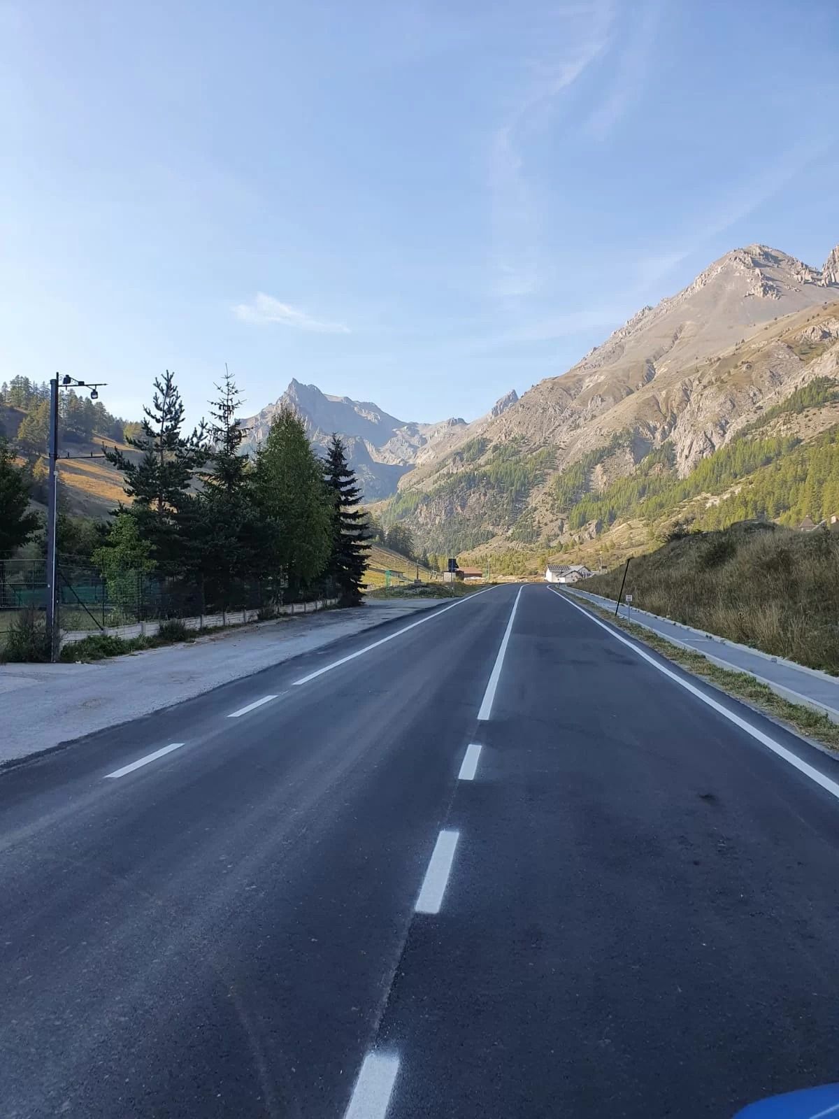Una strada asfaltata rettilinea si addentra in un paesaggio montuoso sotto un cielo limpido e azzurro, punteggiato da pini sparsi.