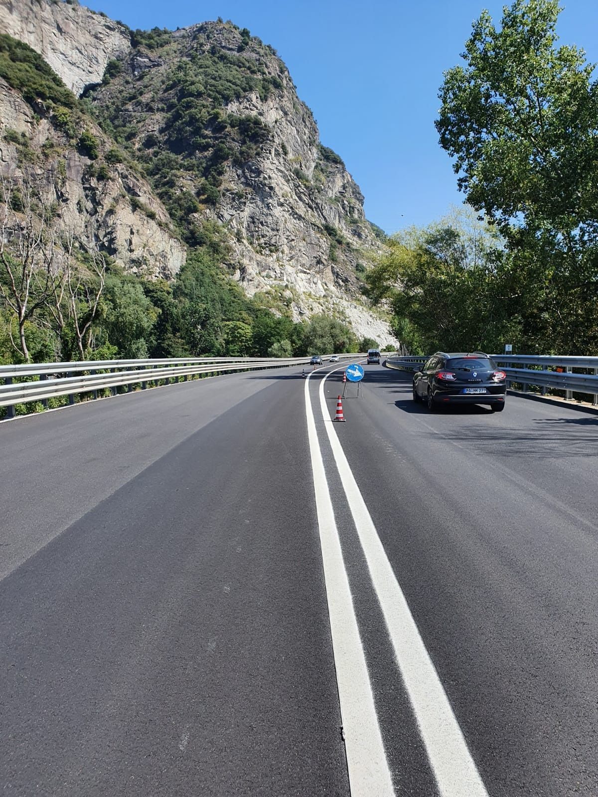 Una strada serpeggia attraverso un passo di montagna sotto un cielo azzurro e limpido, un'auto che procede davanti e dei coni stradali che delimitano la corsia.