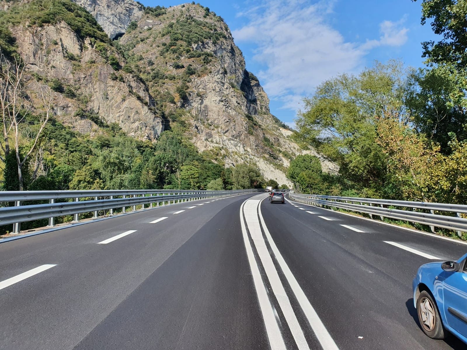 Un'autostrada a più corsie si snoda attraverso un paesaggio montuoso e alberato, con un'auto blu in primo piano.