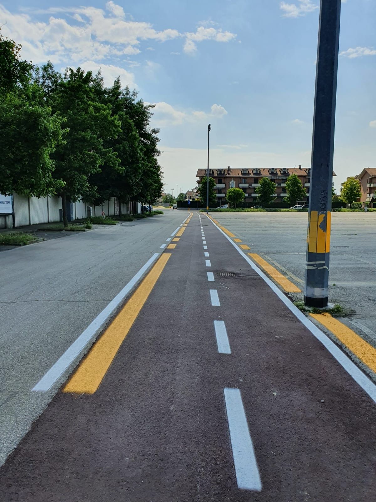 Una pista ciclabile asfaltata, di colore marrone rossastro, con linee tratteggiate bianche, che si snoda tra una strada grigia e un parcheggio.