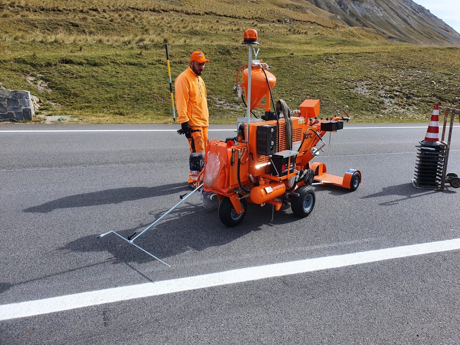 Un operaio con indosso una tuta di sicurezza arancione brillante aziona una macchina per la segnaletica stradale su una strada asfaltata con un pendio erboso.
