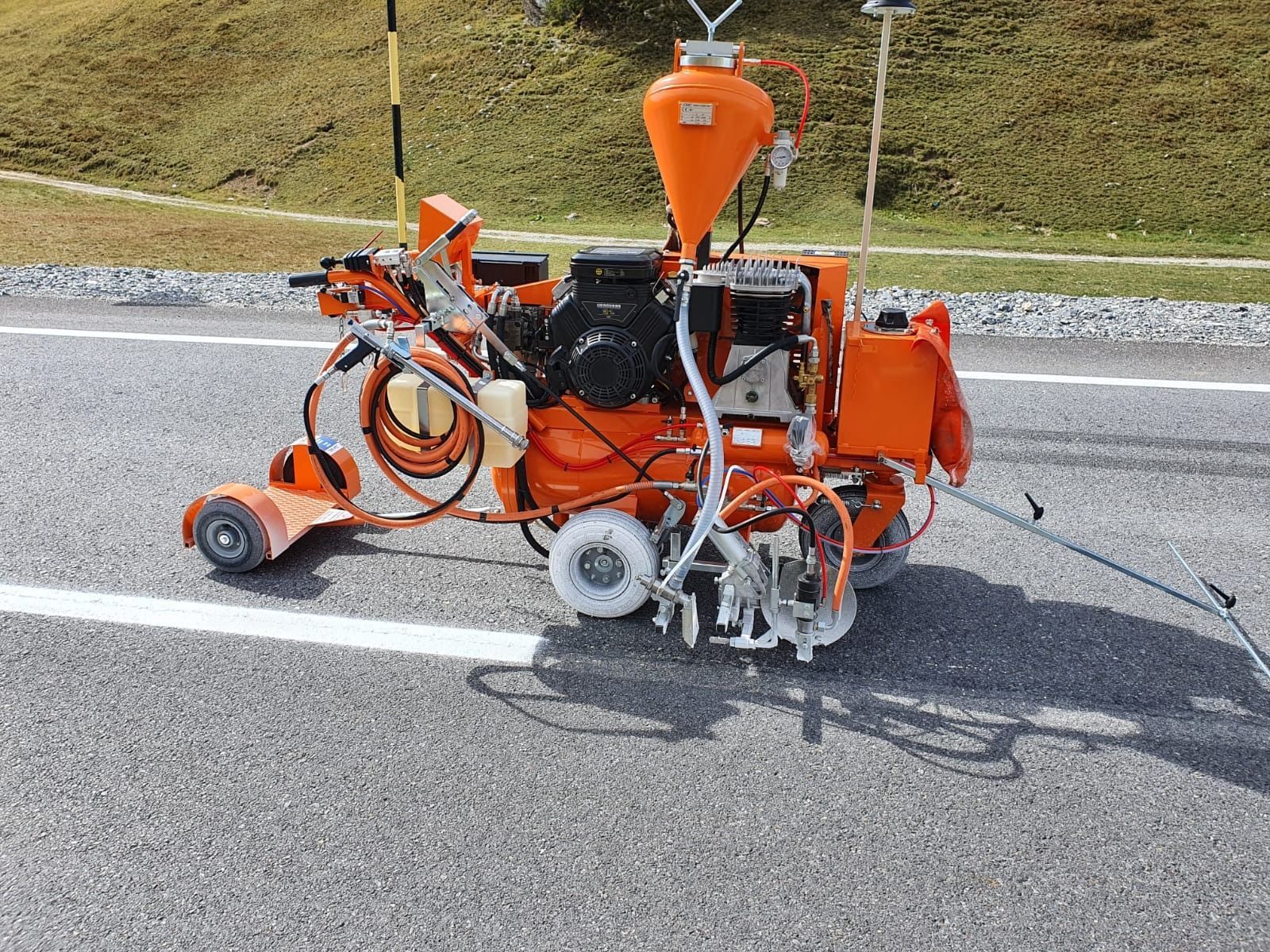 Una macchina segnaletica stradale arancione, semovente, traccia una linea bianca su una strada asfaltata.