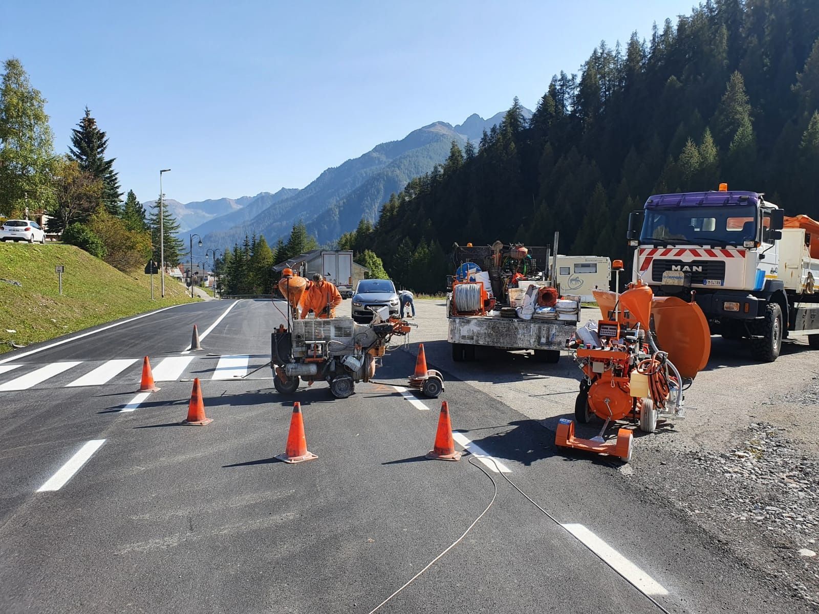 Operai con indosso tute arancioni azionano macchinari per dipingere strisce bianche su una strada vicino a una catena montuosa.