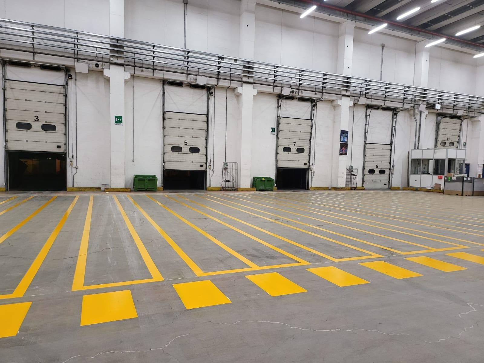 Interno di un magazzino con diverse baie di carico e strisce di sicurezza gialle dipinte sul pavimento di cemento grigio.