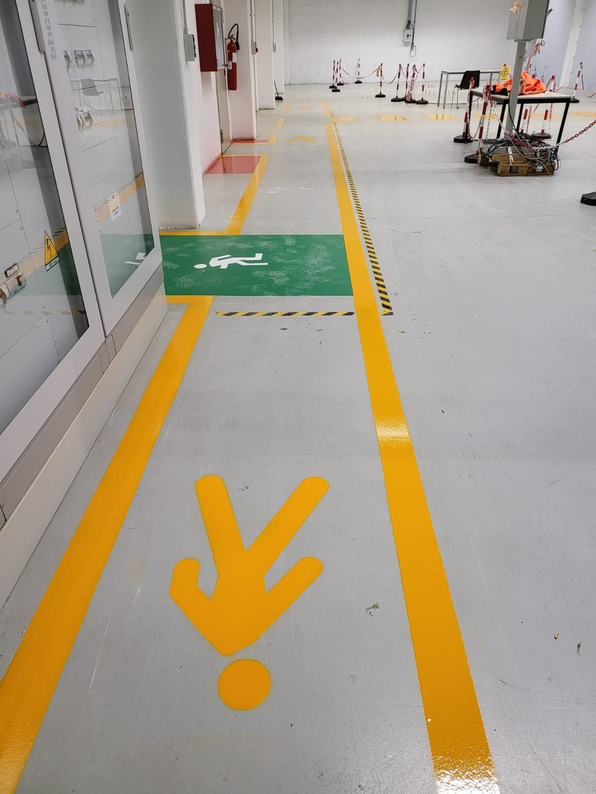 Un percorso pedonale segnalato da linee gialle e dall'icona di una persona su pavimento grigio, che conduce a una zona verde accessibile.