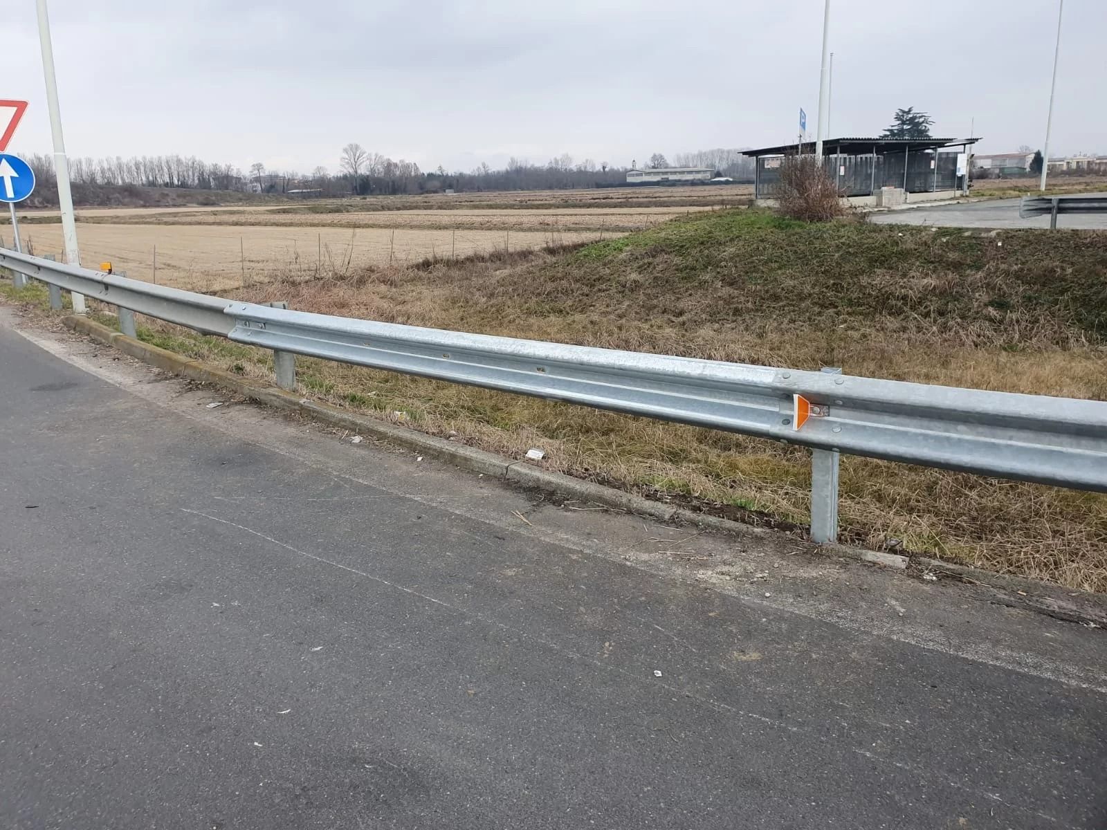 Una barriera metallica corre lungo il bordo di una strada asfaltata accanto a un prato, con una pensilina dell'autobus in lontananza.
