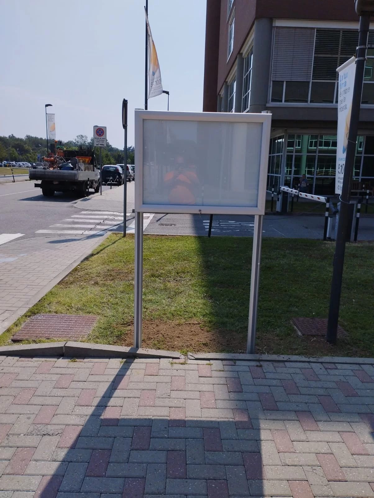 Un pannello espositivo rettangolare vuoto, sorretto da due pali d'argento, si erge su un prato accanto a una strada asfaltata e a un edificio.