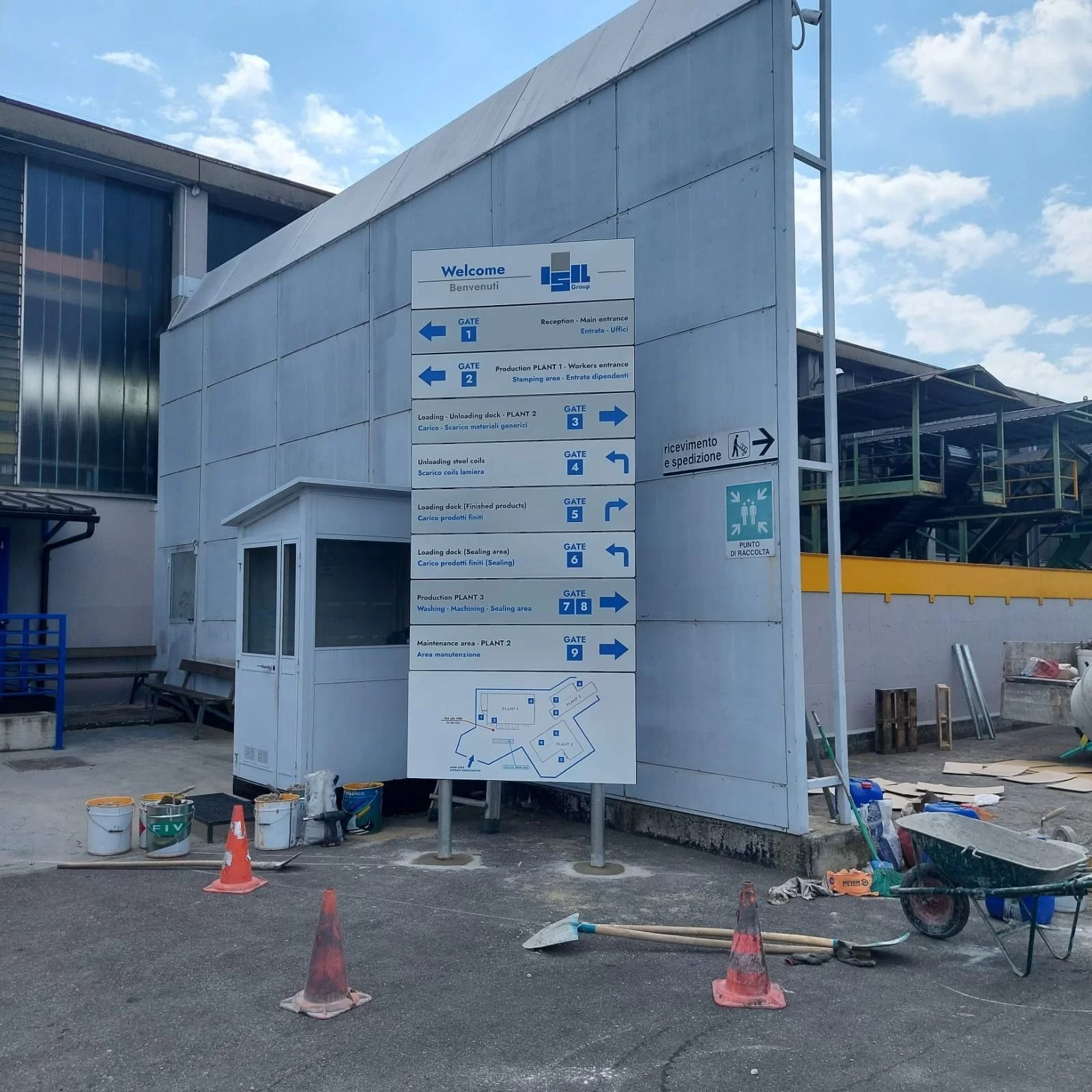 Un cartello direzionale si erge davanti a un edificio bianco sotto un cielo azzurro e nuvoloso, con detriti di costruzione a terra.