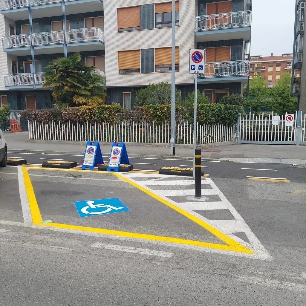 Posto auto riservato ai disabili, segnalato con linee gialle e il simbolo blu di una sedia a rotelle sul marciapiede di fronte all'edificio.