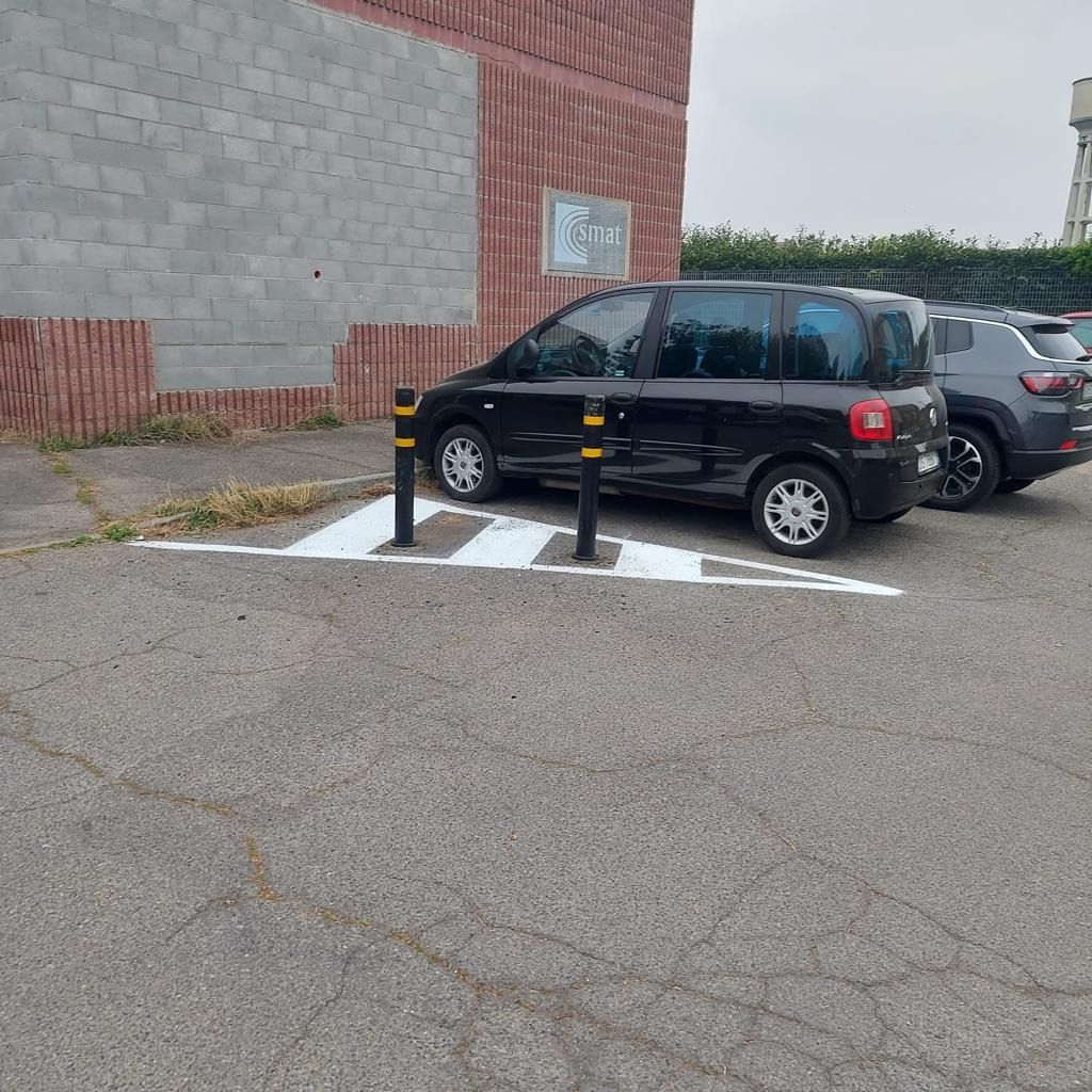 Un'auto nera parcheggiata accanto a un'isola spartitraffico triangolare dipinta di bianco, delimitata da due dissuasori, su un piazzale asfaltato di fronte a un muro di mattoni.