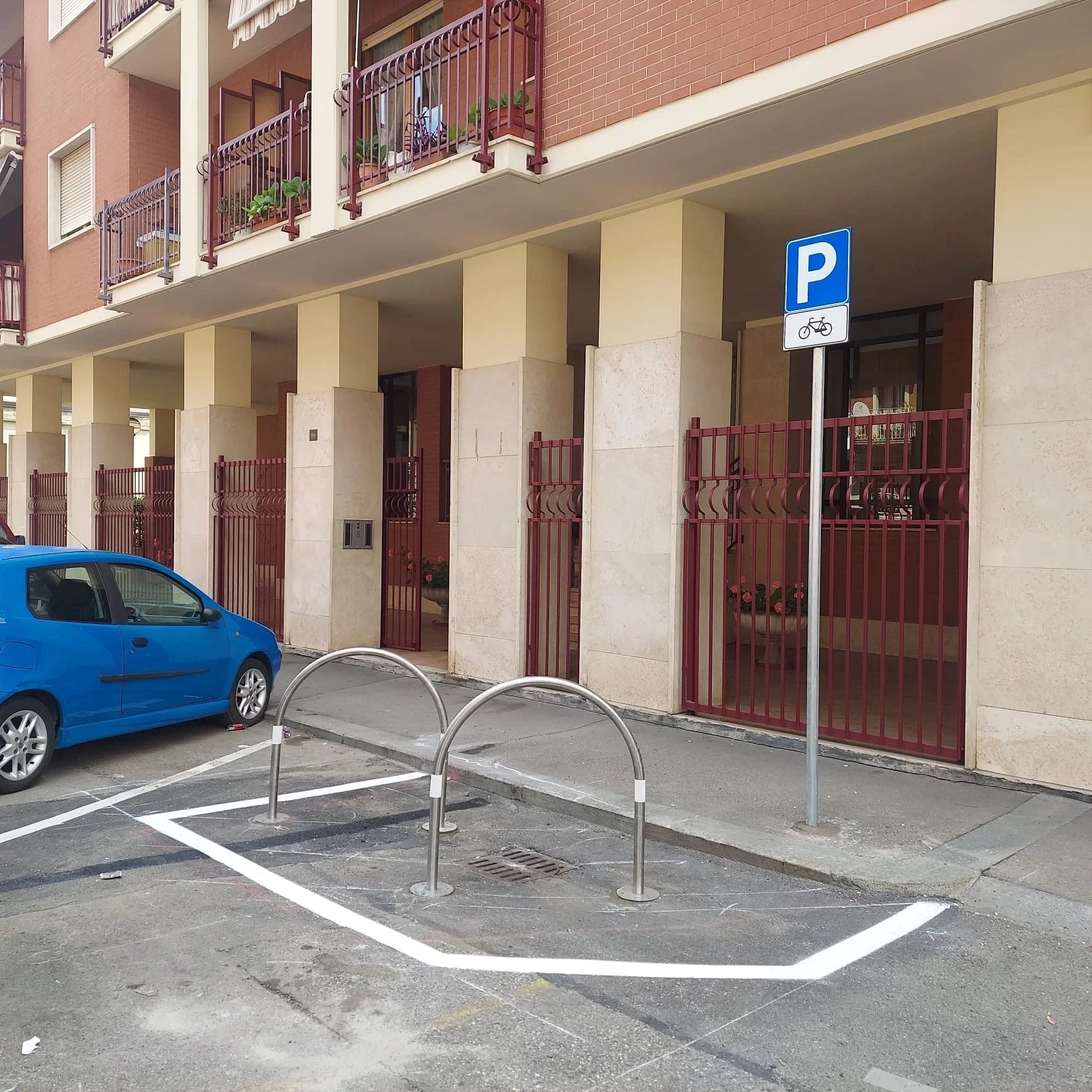 Un'auto blu parcheggiata vicino a un'area di parcheggio per biciclette segnalata, con due rastrelliere metalliche e un cartello di parcheggio per biciclette sul marciapiede.