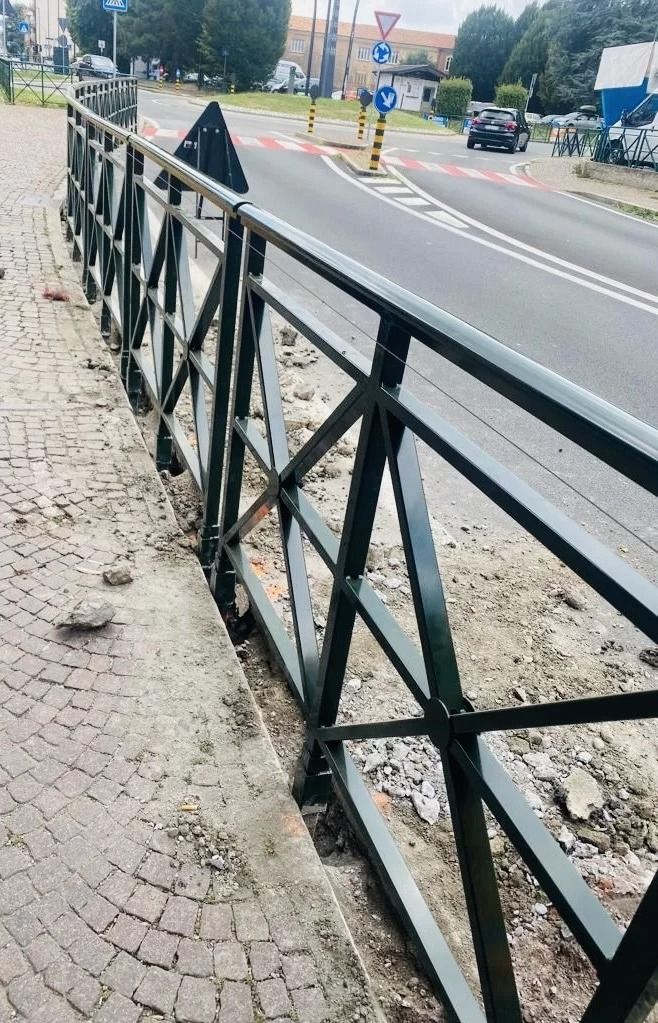 Una ringhiera pedonale in metallo nero costeggia un marciapiede lastricato, accanto a una strada asfaltata, con traffico e segnaletica in lontananza.