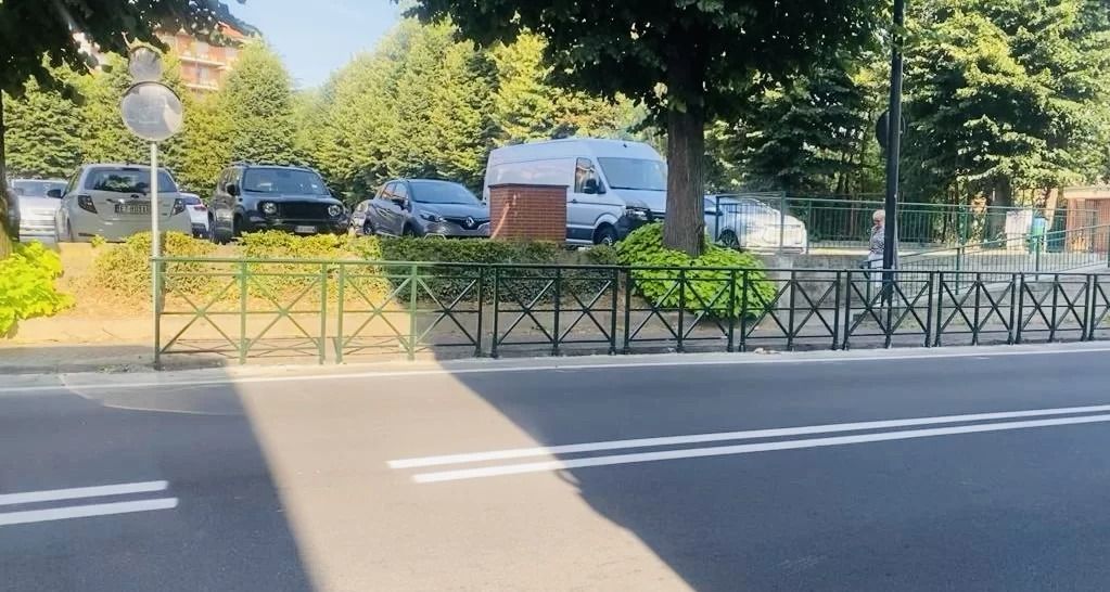 Una scena di strada con una carreggiata asfaltata, una recinzione metallica pedonale e un parcheggio con diverse auto parcheggiate sotto gli alberi.