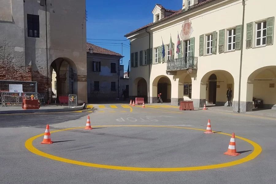 Una segnaletica circolare gialla con coni stradali su una piazza pavimentata, di fronte a un edificio con passaggi ad arco.