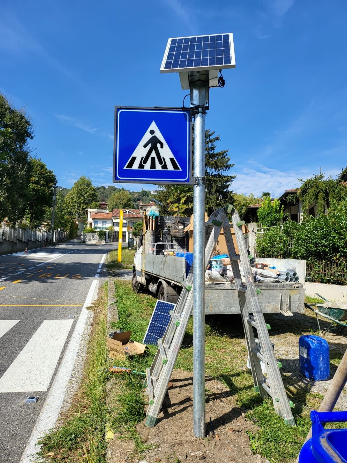 Un segnale pedonale blu con un pannello solare sulla parte superiore, viene installato a bordo strada con una scala e un camion di servizio.