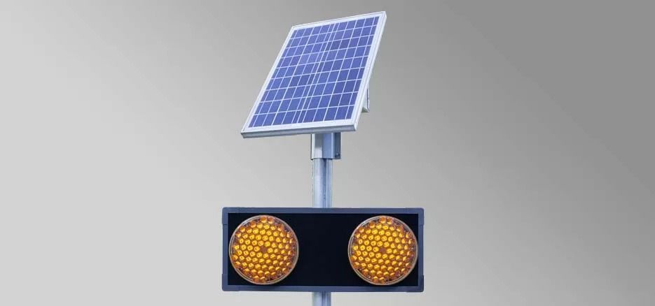 Un semaforo a energia solare con due luci rotonde lampeggianti di colore ambra, montate su un palo grigio su sfondo grigio.