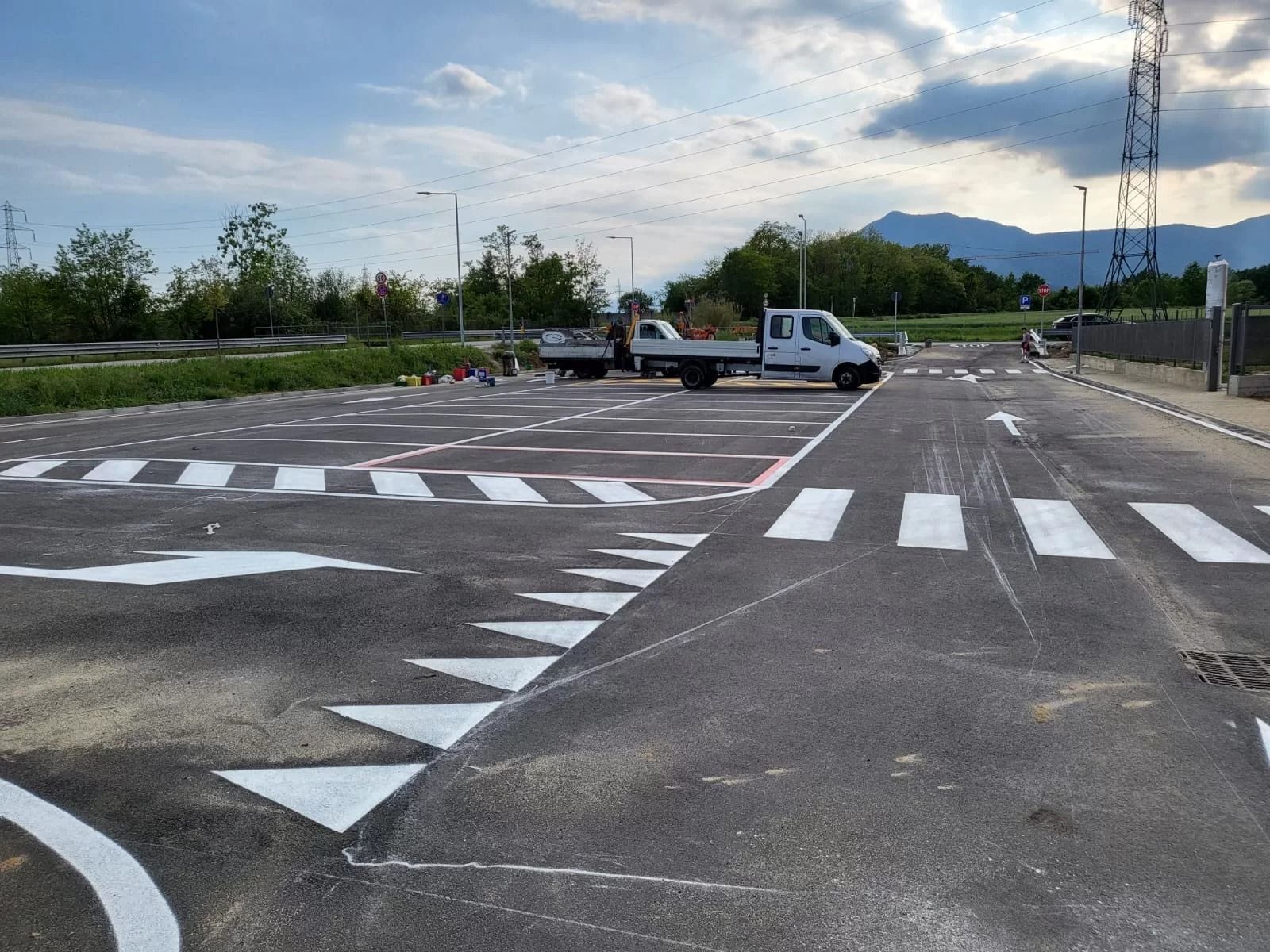 Un parcheggio asfaltato sotto un cielo nuvoloso, con segnaletica stradale bianca, attraversamenti pedonali e un camion bianco.