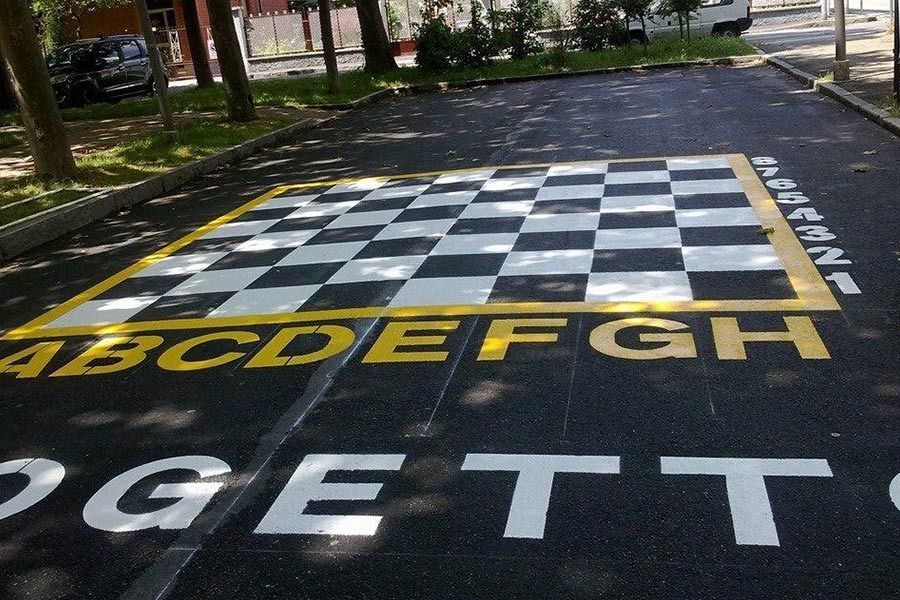 Una grande scacchiera dipinta sull'asfalto, contrassegnata dalle lettere AH e dai numeri da 1 a 8, situata in un'area esterna.