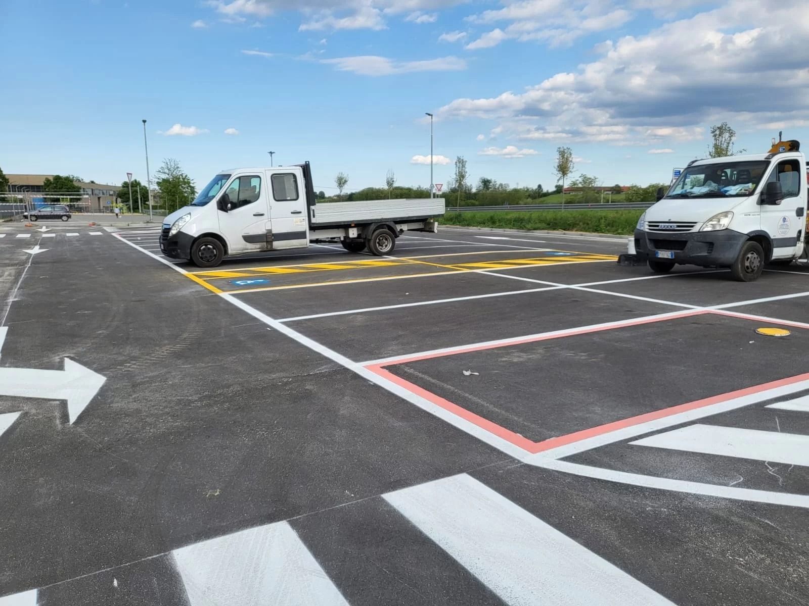 Due furgoni bianchi di servizio parcheggiati in un parcheggio esterno appena asfaltato e segnalato, sotto un cielo azzurro e soleggiato.