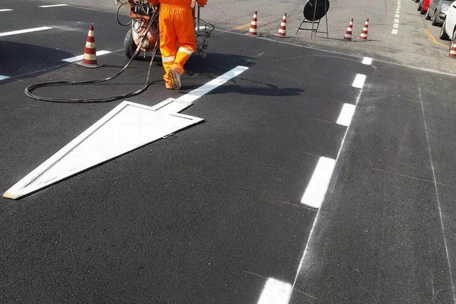 Un operaio con indosso una tuta arancione brillante spruzza una freccia direzionale bianca su una strada di asfalto nero appena asfaltata.