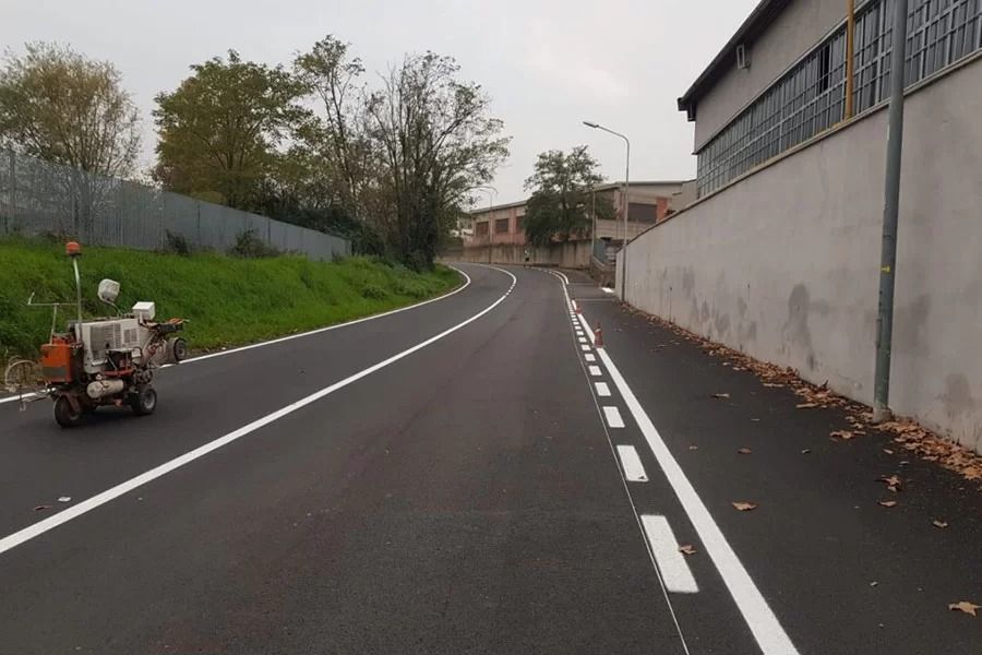 Una macchina specializzata traccia linee bianche su una strada appena asfaltata, delimitata da un muro grigio e da un terrapieno erboso.