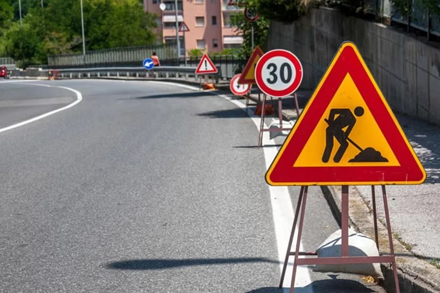 Un cartello giallo di lavori stradali con bordo rosso si erge su una strada, con un cartello del limite di velocità e altri segnali di pericolo alle sue spalle.