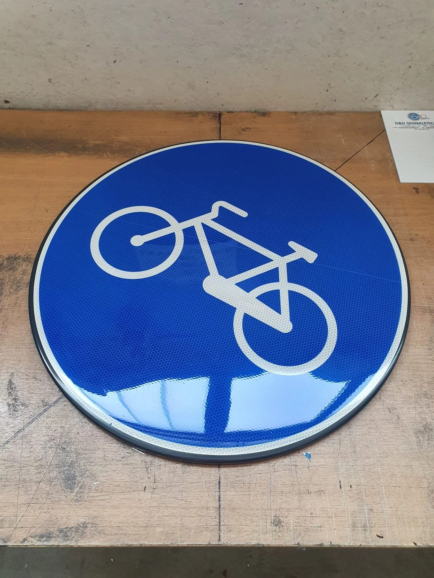 Un segnale stradale circolare blu con il simbolo di una bicicletta bianca, che indica un percorso ciclabile, posizionato su una superficie di legno.