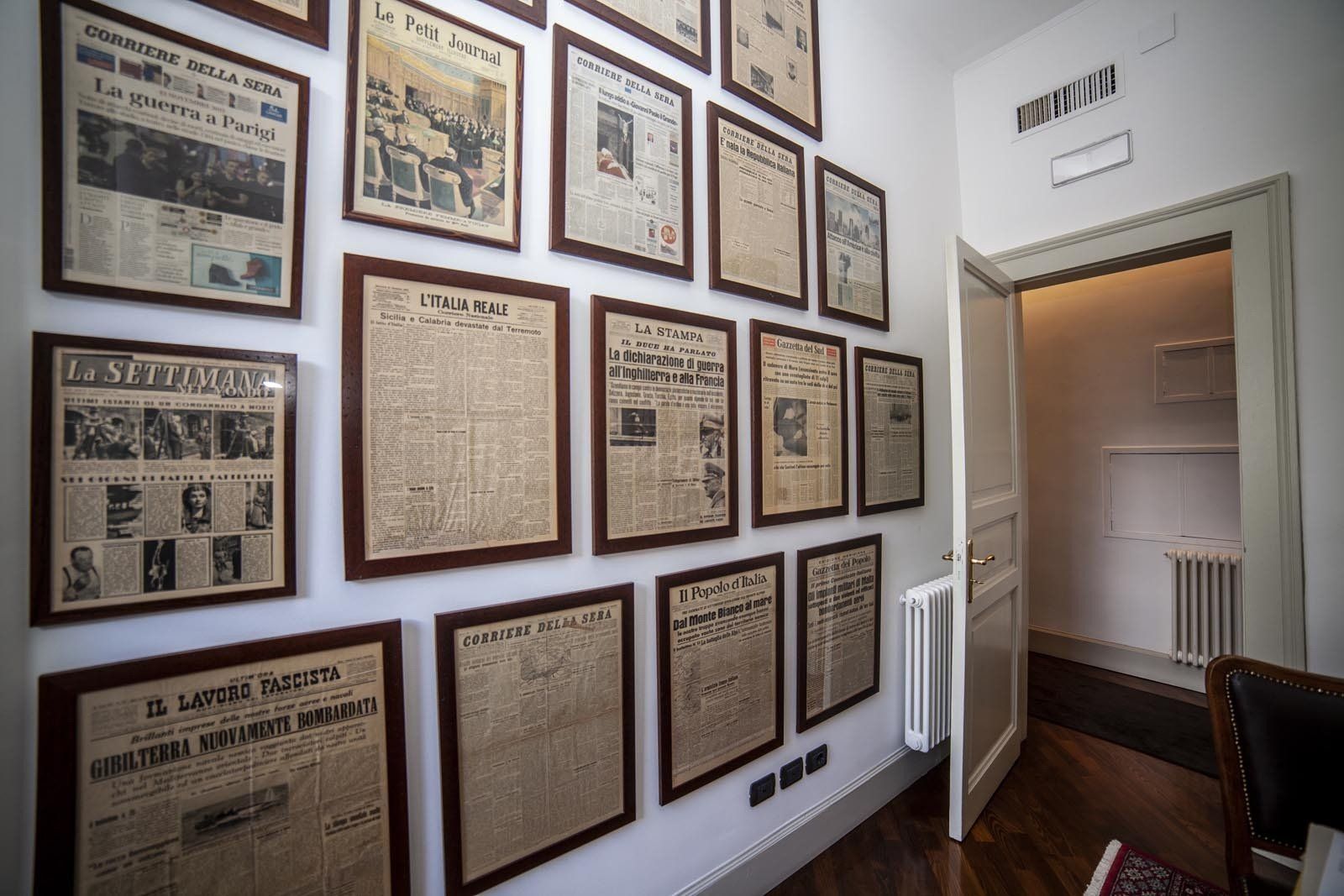 Parete con giornali d'epoca incorniciati. Porta aperta sulla destra. Pavimento in legno e pareti bianche.