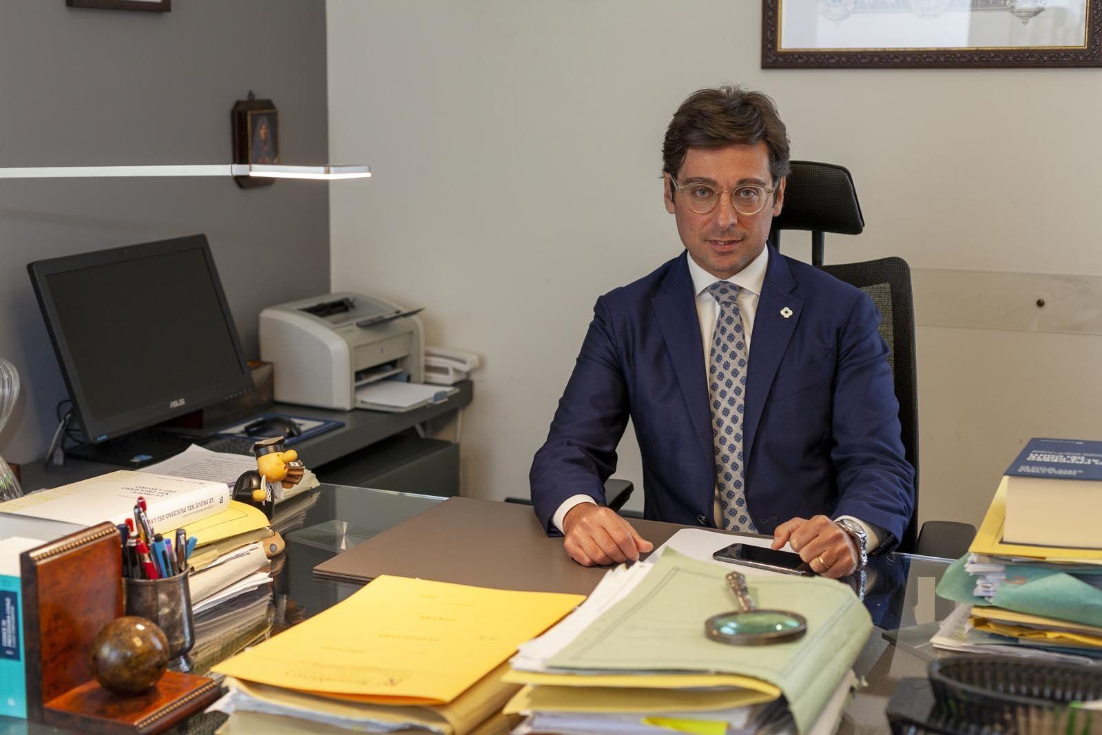 Uomo in giacca e cravatta seduto alla scrivania, guarda la telecamera. Ambiente d'ufficio con documenti e computer.