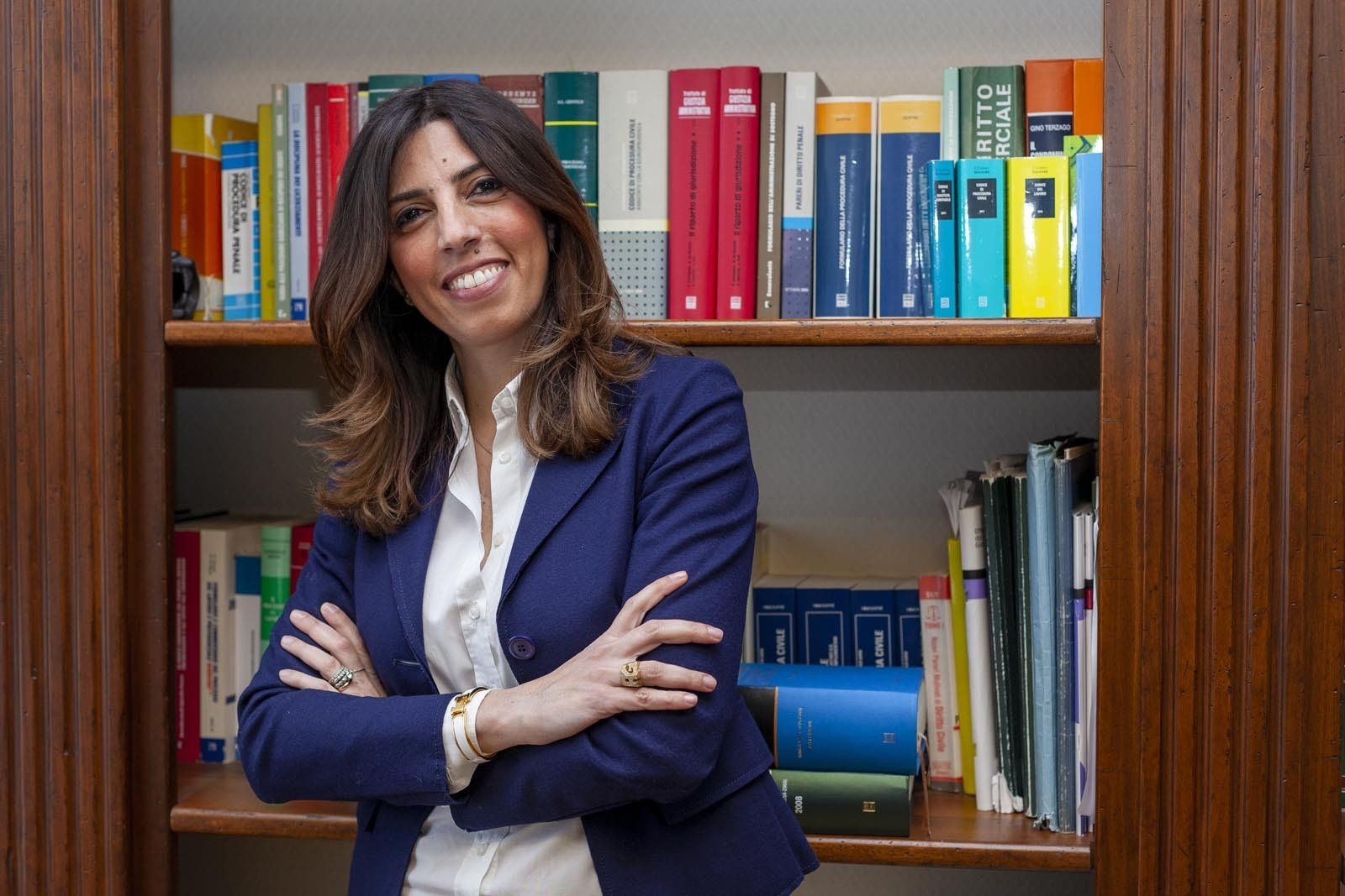 Donna con i capelli castani in giacca sportiva, in piedi vicino a una libreria, sorridente e con le braccia incrociate.