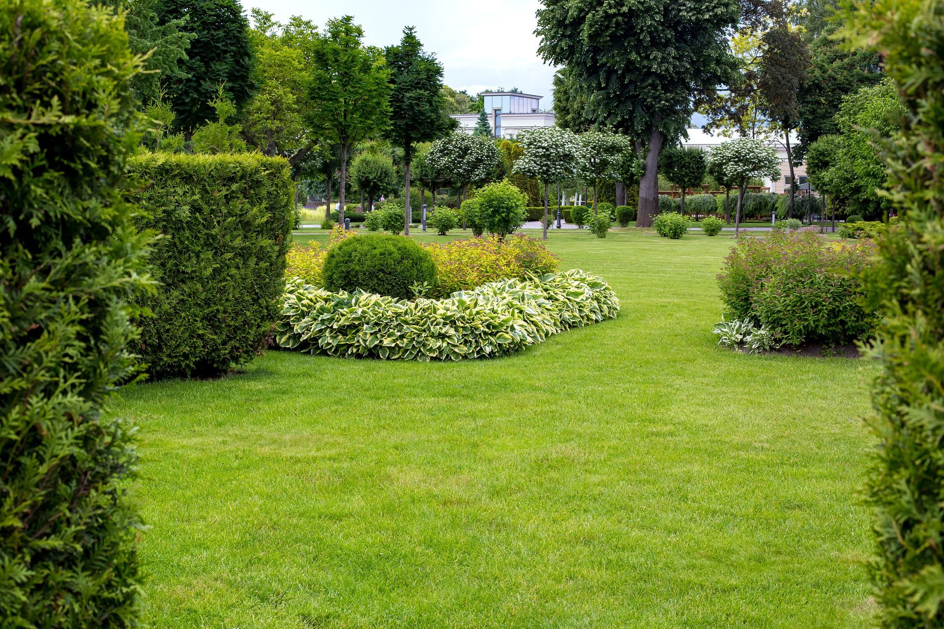 A lush green park featuring manicured hedges, a circular flower bed, and various trees under a cloudy sky.