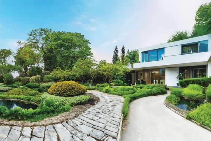 A modern white building overlooks a lush garden with a stone path, green hedges, and a small pond under a clear blue sky.