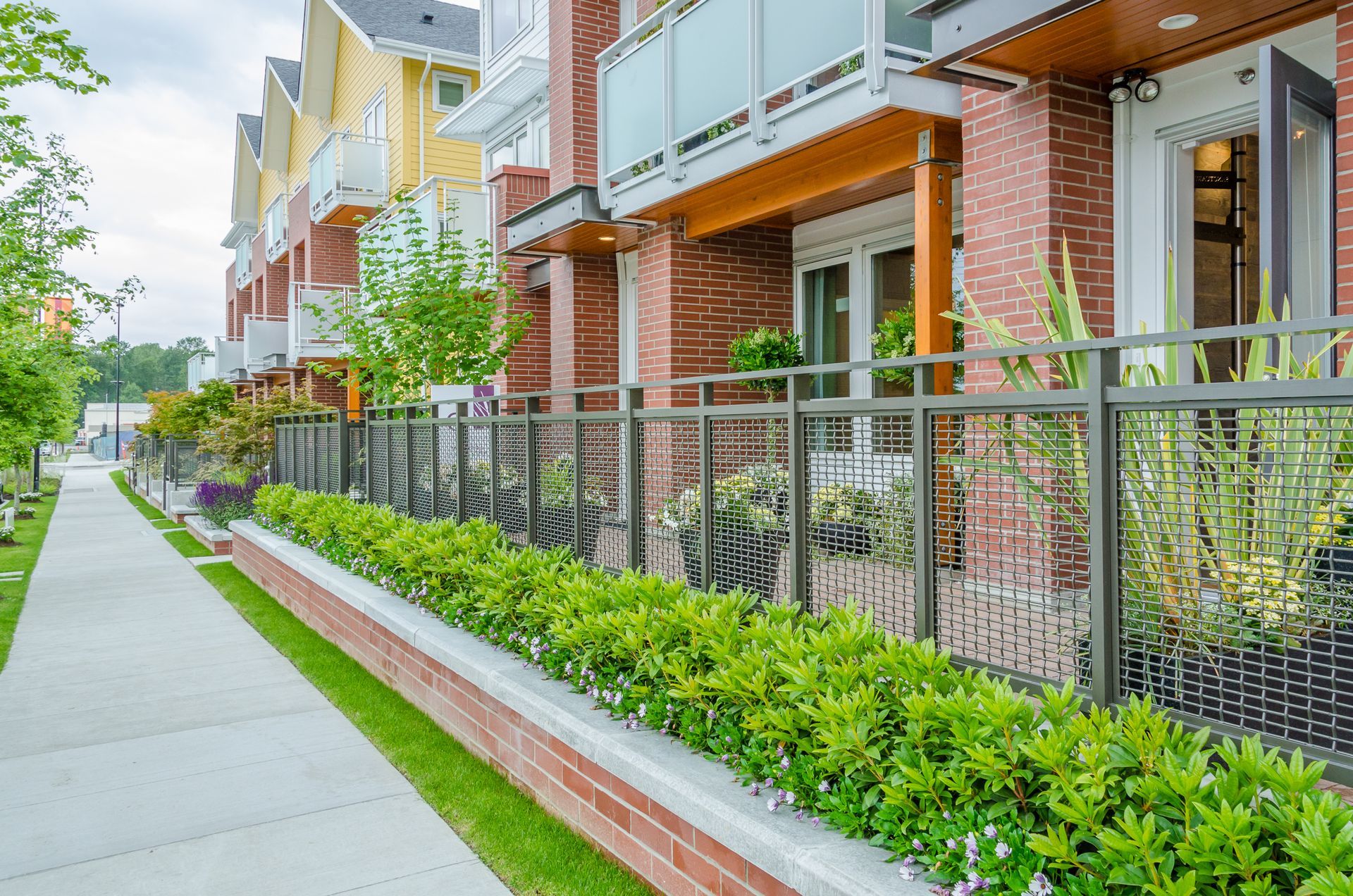 Row of townhomes with brick facades, balconies, and a sidewalk lined with greenery.