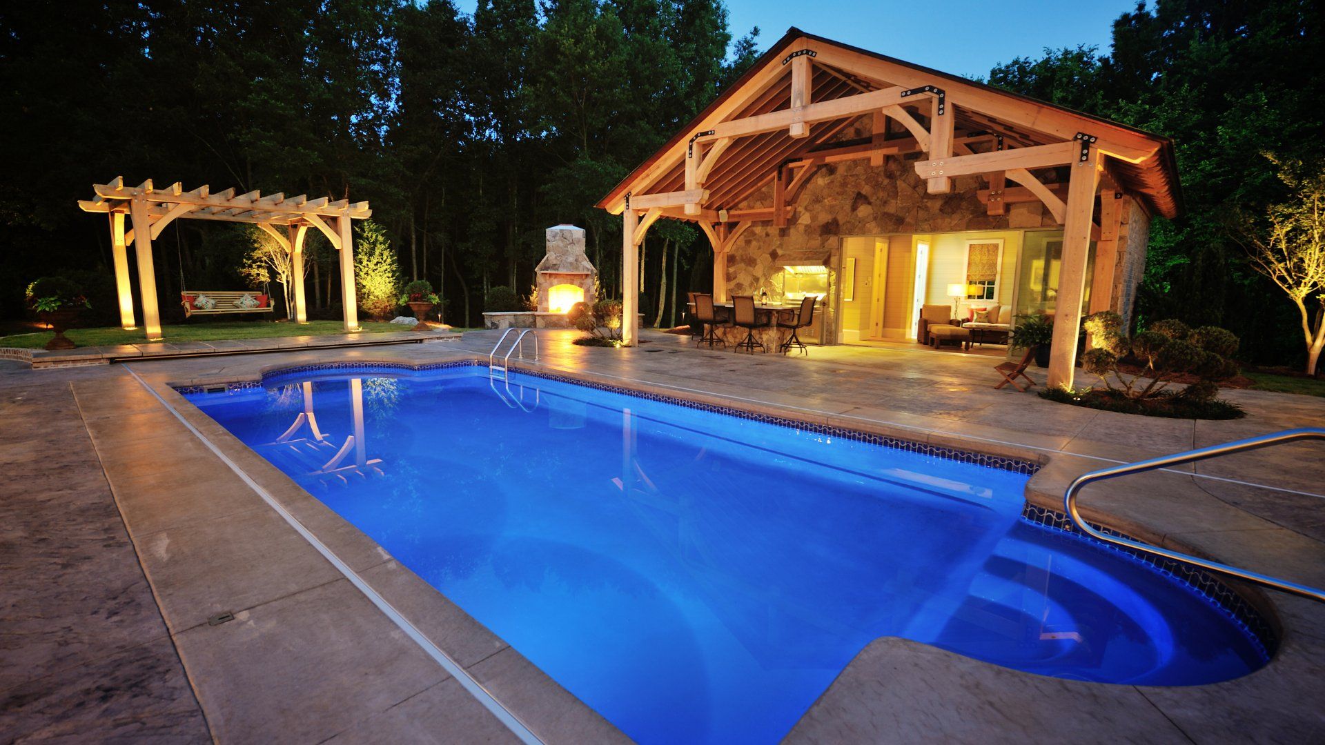 Backyard pool at night.  The pool is lit up.  The backyard also features a covered patio and pergola.  The deck around the pool is stamped concrete.