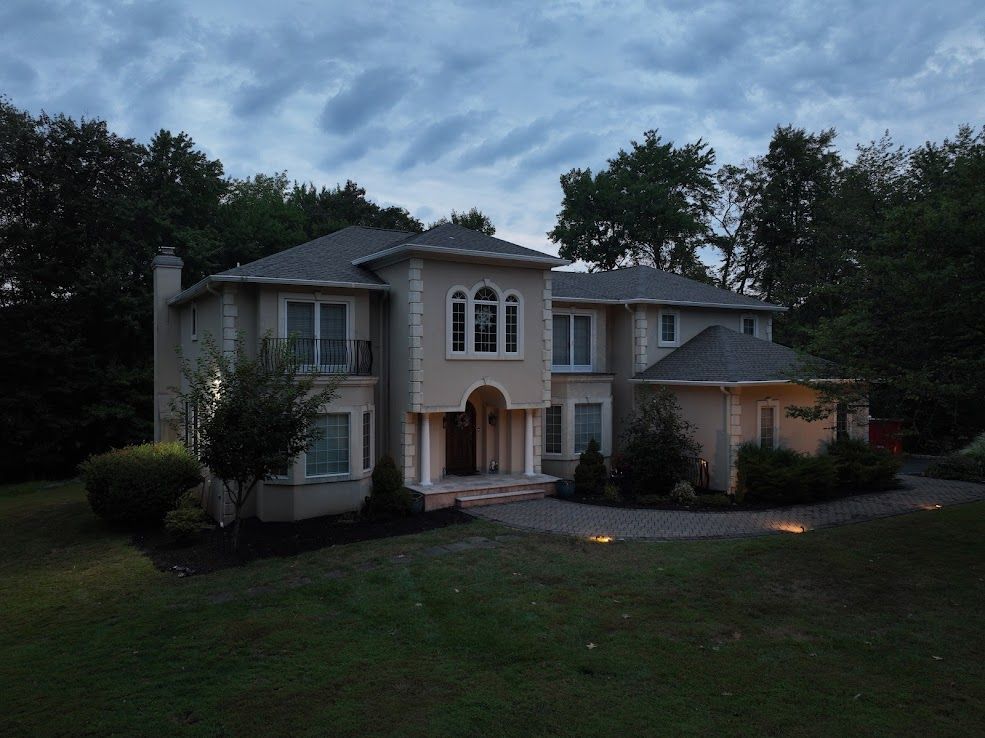 Large two-story house with tan stucco exterior, multiple windows, and a circular driveway, under a cloudy evening sky.