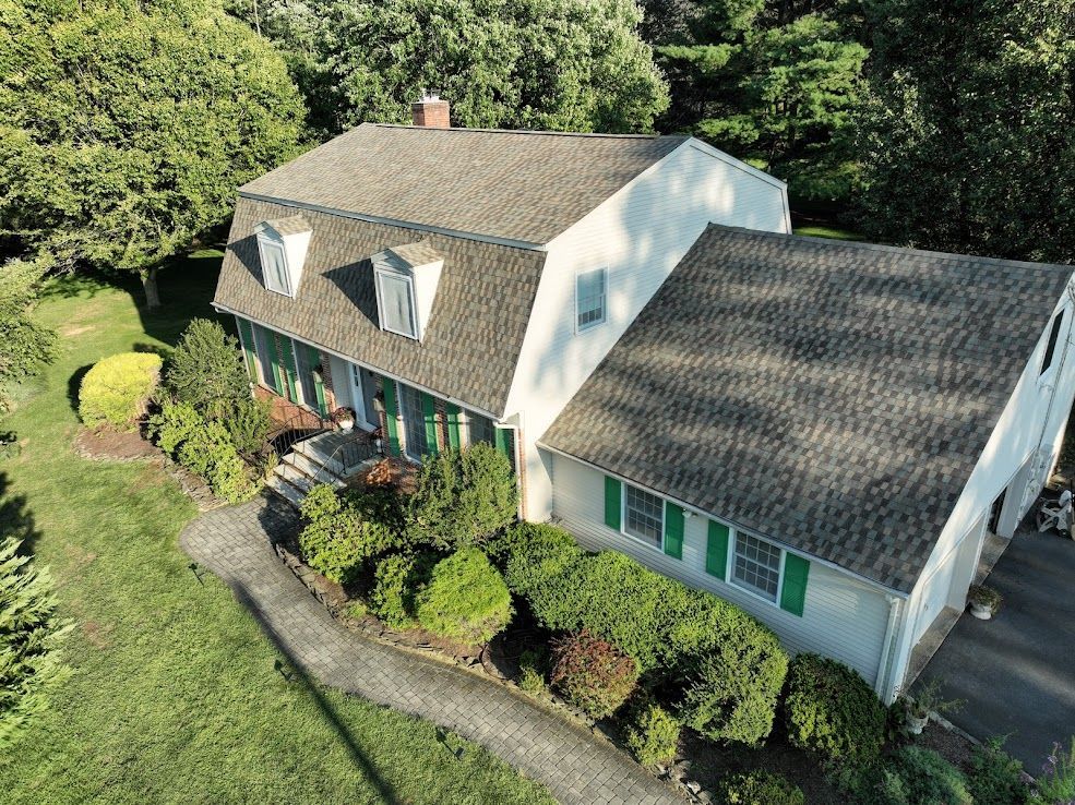 Two-story white house with green shutters, dormers, and asphalt roof, surrounded by landscaping and trees.