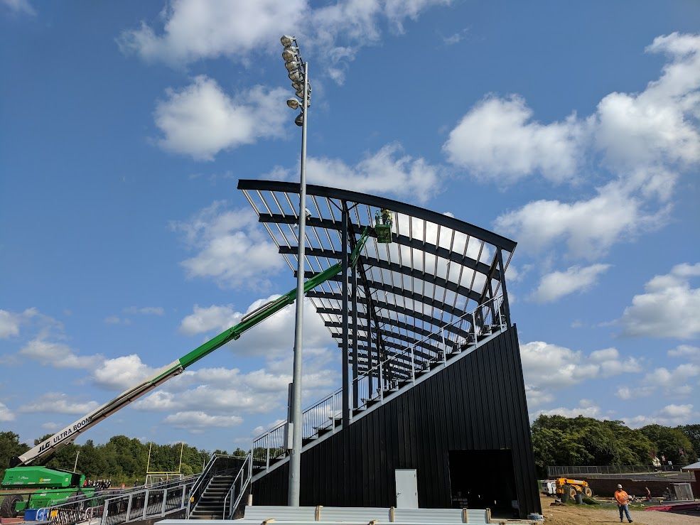 Bleacher seating under construction with lift and light tower against blue sky.
