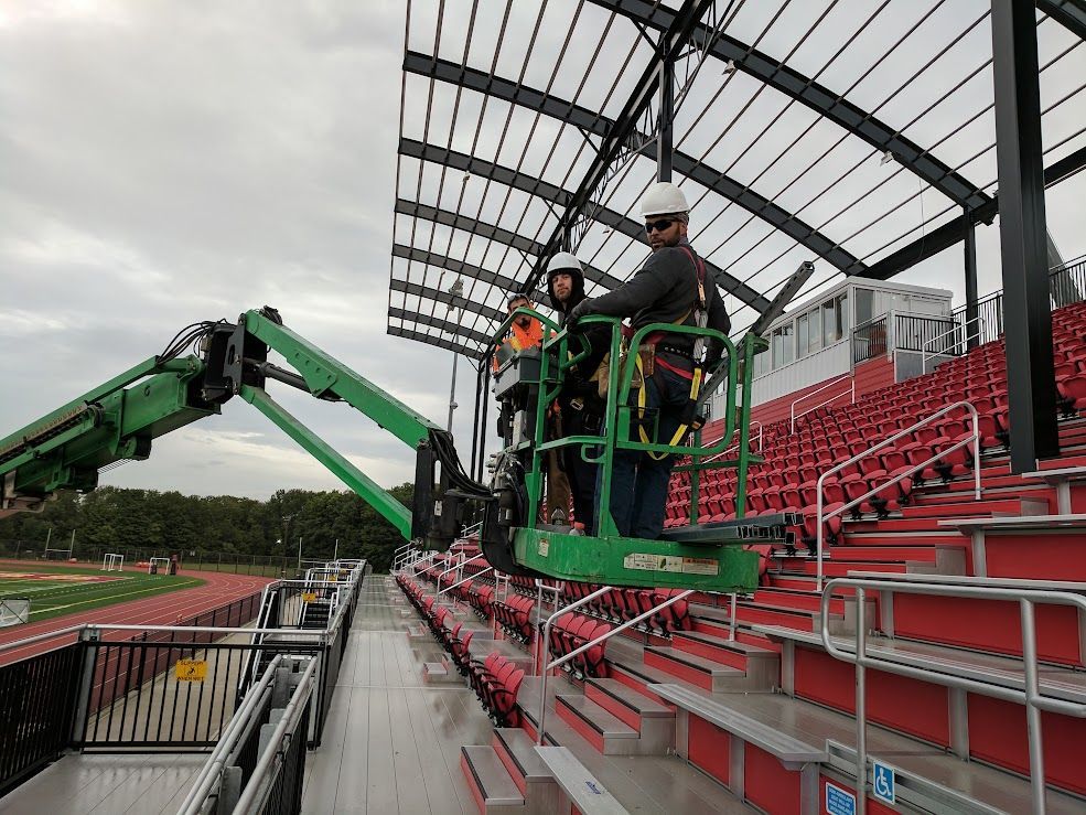 Workers in a lift installing roofing at a stadium with red seating. Cloudy day.