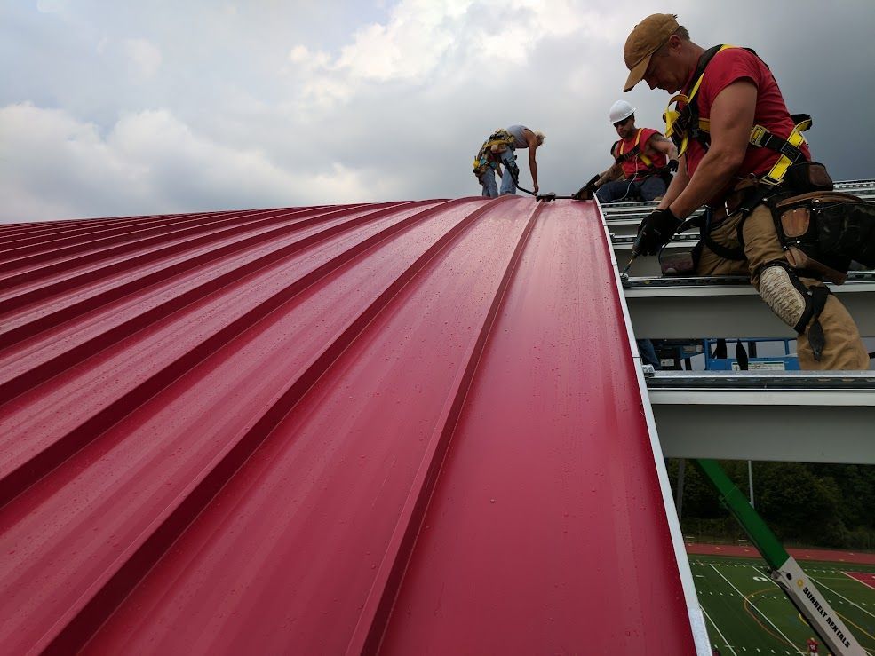 Roofers installing red metal roofing on a curved roof structure, under cloudy skies.