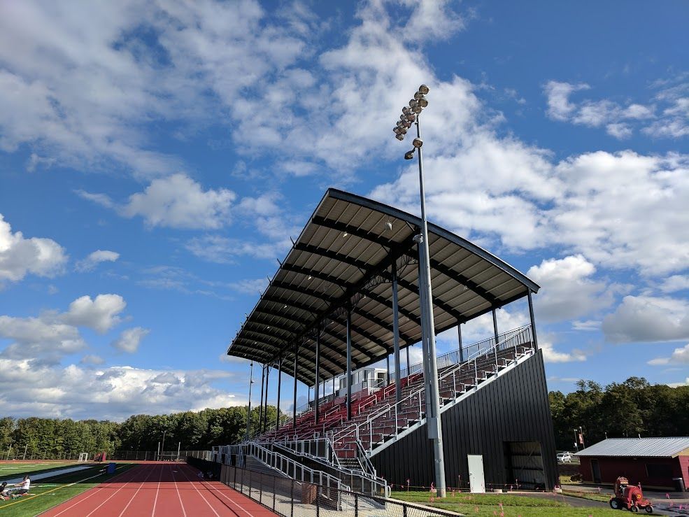 Bleachers at a track and field stadium, under a blue sky with clouds. Red track in foreground.