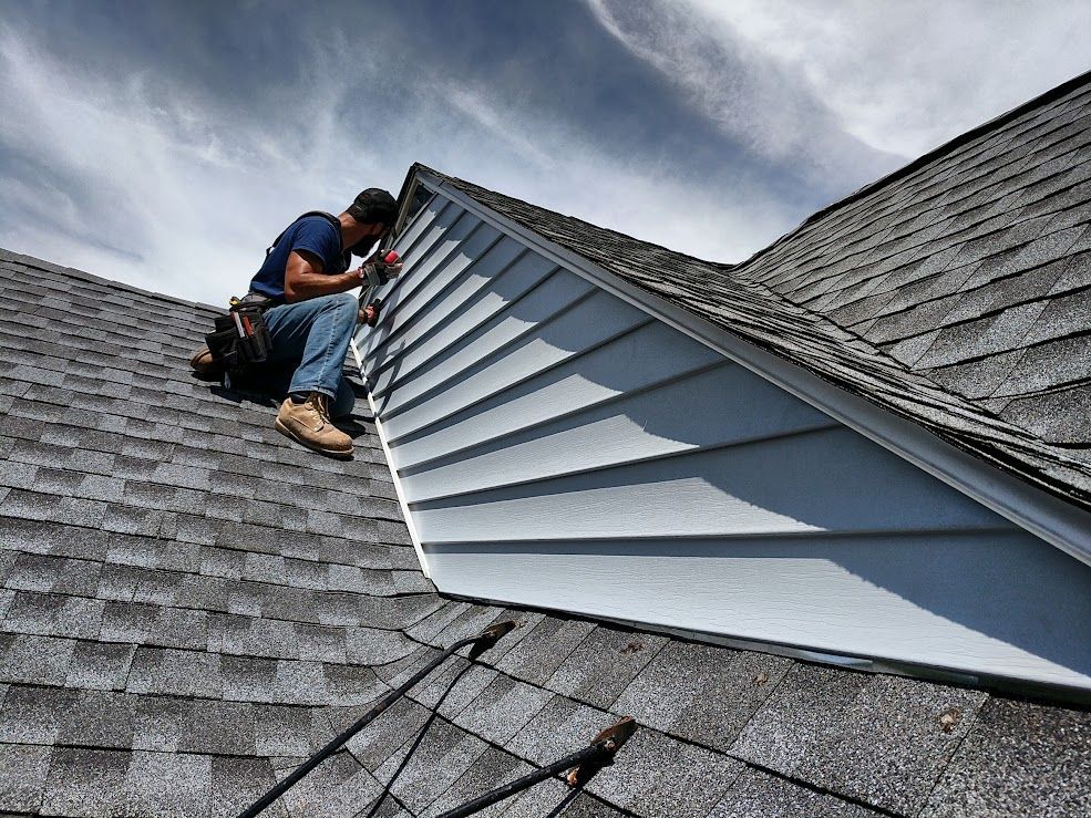 Man on a roof installing siding on a gable. Blue sky, gray shingles, and tan work boots.