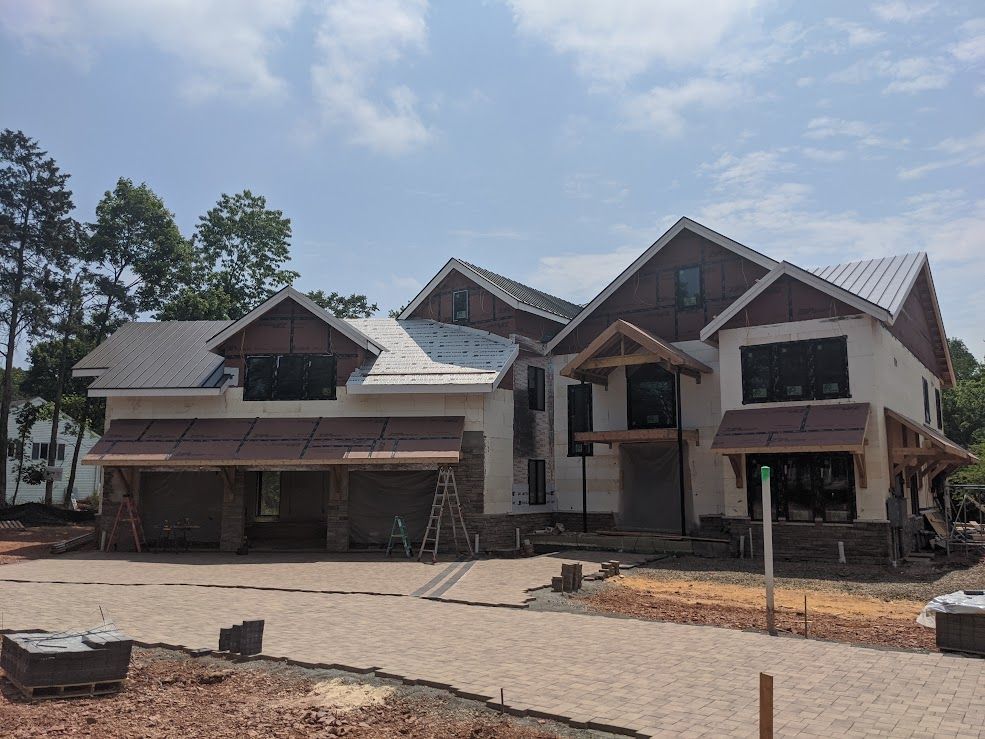 Two-story house under construction with stone and white stucco exterior, grey metal roof, and exposed wood trim.