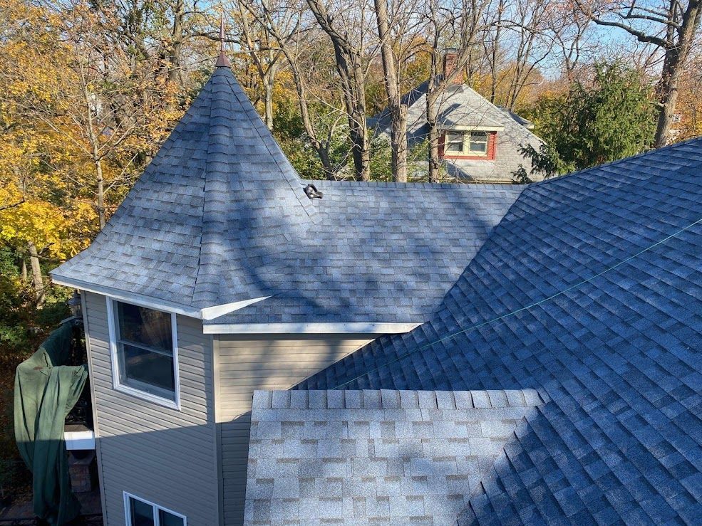 Blue shingled roofs of a house with a turret, tan siding, and trees in the background.