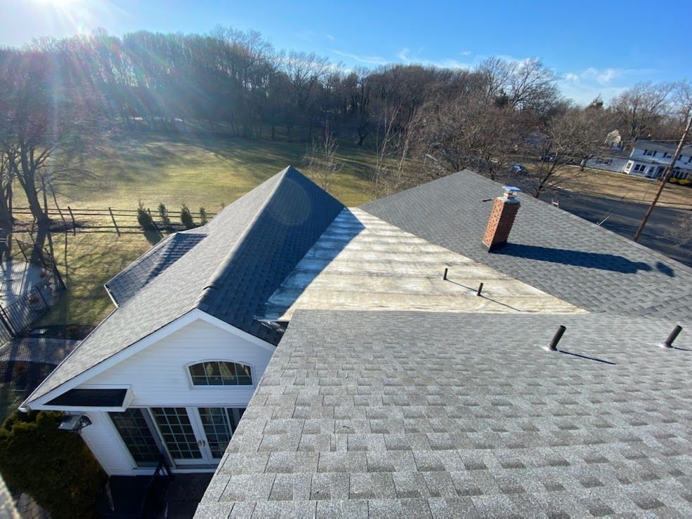 Overhead view of a house roof with light grey shingles, a chimney, and a sunny, tree-lined background.
