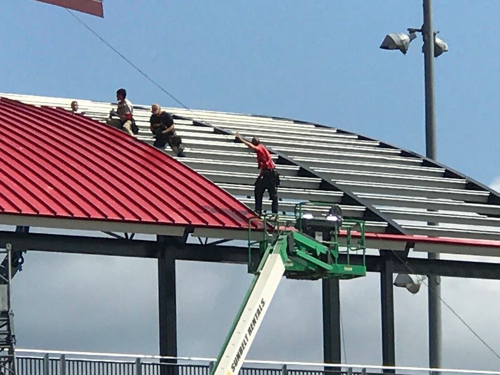 Workers installing metal roofing on a stadium under construction; one on a lift reaching.