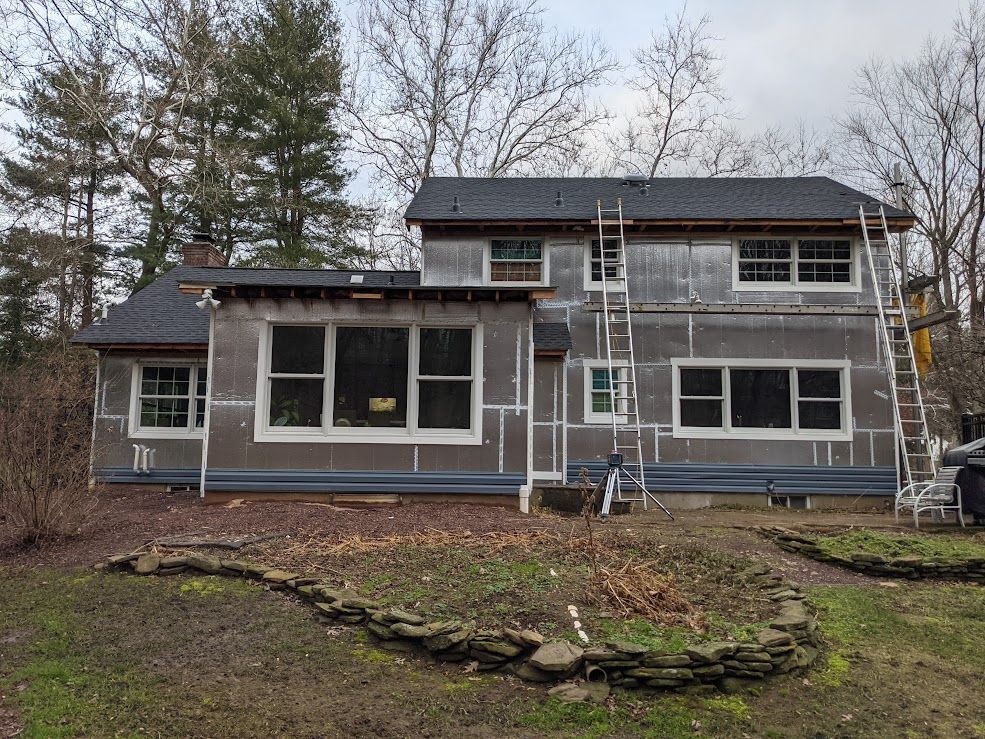 House exterior under construction, covered with insulation, ladders, windows, and surrounding vegetation.